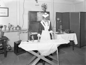 Black and white photo from 1913/14 of a young white woman dressed in a maid’s uniform standing in an appliance shop, laying an apron over an ironing board.
