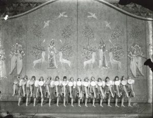 A line of showgirls kicking their legs in unison in front of an Urban-designed backdrop.