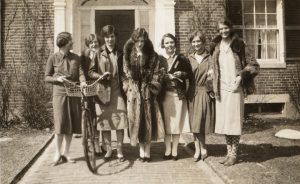 A group of young white women stand in front of a building, smiling at the camera. They have bobbed hair and wear casual dresses. The woman to the very left holds onto a bicycle.