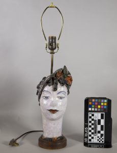 A ceramic head of a woman, painted in white, red, and brown. She has dark curls and roses peeking out from beneath a small brown hat that is tilted to cover her right ear. A lamp socket and an electrical cord are sticking out of the head.