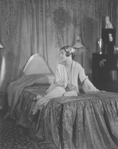 Black and white photo of Elaine Wormser seated casually on her bed in her bedroom in light-colored pajamas, looking away from the camera.