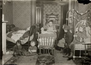 A white family of a woman and five children, and a baby crib, sit in a small and dingy room.