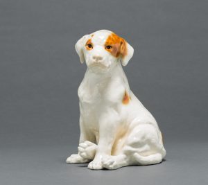 Seated white dog with orange spots around its eyes.