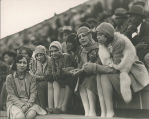 Six young women sit in a crowded stadium, with bobbed hair, cloches, raised hemlines, and scarves. Four of the six women look directly at the camera, the others look towards what is happening on the field.