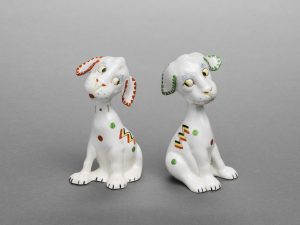 Two white ceramic dogs, painted with orange and green dots, sitting with their heads cocked to the side.