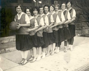 A line of young white women wearing matching uniforms and holding each other by the waist. The woman at the front of the line holds a basketball.