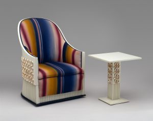 Ivory-colored curved-back chair with red, blue, and yellow striped upholstery next to an ivory-colored table with gold embellishments.