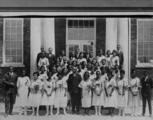 A large group of young Black women in white dresses and a few Black men in suits stand in rows on the steps of a building. Each woman has a flower pinned to her dress.