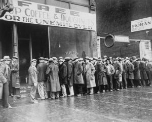 A long line of men stand outside of a shop with the sign, “Free Soup Coffee Donuts for the Unemployed.”