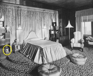 A black and white photo of Elaine Wormser’s room, showing her bed and two bookcases. A small ceramic dalmatian stands on the lower shelf of the bookcase on the left. It is circled in red in the picture.