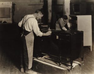 Photograph from 1923 of two white men, in long aprons, polishing large pieces of furniture.