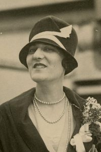 Photo of a white woman wearing a black coat, a pearl necklace, and a black cloche with a white zigzag ribbon, smiling at the camera.