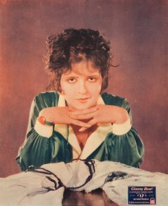 A young white woman, dressed in green and white, with brown eyeshadow and short curly hair folds her hands under her chin and looks directly at us.