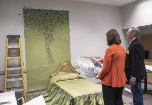 A woman and a man, with their backs to the camera, compare Elaine Wormser’s bed and a reproduction of her curtains to an enlarged archival photograph of her room held by the woman.