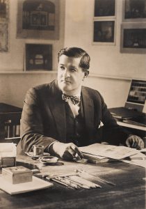 Joseph Urban seated at his desk, looking away from the camera. Framed pictures hang on the wall behind him.