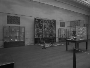Black and white photo from 1926 of a museum gallery filled with cases containing small decorative objects and a large folding screen.