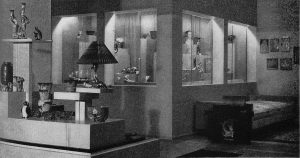 Black and white photo of a room featuring a dramatically lit corner case and shelves with small decorative objects on display.