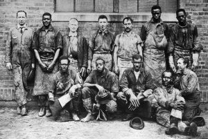 Photograph from circa 1930 of a team of Black and white men dressed in coveralls and aprons, looking directly at us. Seven men stand against a brick wall, while five kneel in front. Their clothes are heavily stained with chemical dyes.