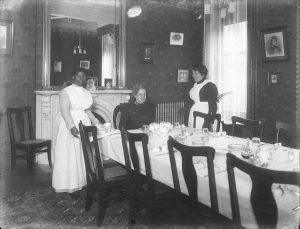 Black and white photo from 1913 of a white woman and a Black woman dressed in maid’s uniforms serving a seated white woman in a dining room.