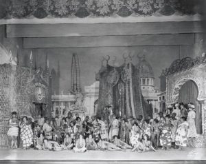 A large cast of characters assembled in front of a highly ornate, Urban-designed set of a city