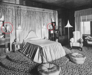 Black and white photo of Elaine Wormser’s bedroom, with her bed in the center and two bookcases to the left and right. On each bookcase is a silver urn shaped like a rocket. The urns are circled in red in the image.