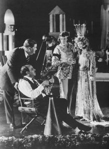 Joseph Urban looking on as a film director speaks with two cast members dressed as a medieval prince and princess.