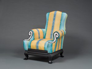 A cushy armchair, low to the ground, with curved sides, blue and yellow striped upholstery, and white spirals on the front of its arms.