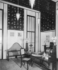 Black and white photo of an interior with dark furniture, dark floral wallpaper, a silver tea service and vase, and triangular hanging lamps.