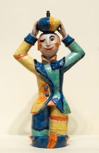 Winking ceramic boy in a colorful suit of blue, pale yellow, teal and orange, holding a ball on top of his head.