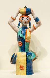 Ceramic girl in a colorful dress of blue, pale yellow, teal and orange, holding a ball on top of her head.