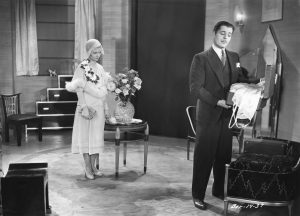 A film still from 1931 depicting a man and woman standing in a circular interior featuring dark, glossy furniture with white detailing.