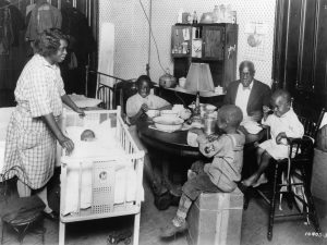 A Black family of a woman, a baby, three young boys, and a man sit and stand in a small room around a table for mealtime. The boys are smiling.