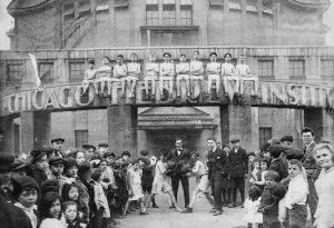 A crowd of young Jewish boys and girls gather around an archway emblazoned with the words “Chicago Hebrew Institute.” A row of boys wearing boxing shorts sit upon the archway, while two boys box below.