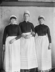 Black and white photo from 1888 of three white women dressed in maid’s uniforms standing closely together, looking directly at us.