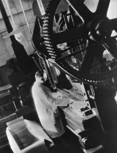A machine with large gears is operated by a white man wearing a coat, looking down at a sheet of plastic.