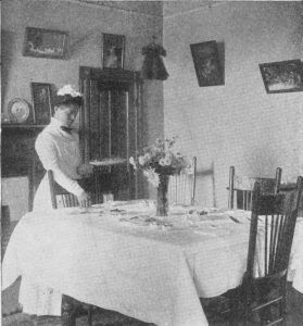 Black and white photo from 1910 of a young Black woman dressed in a maid’s uniform, looking down as she places cutlery on a dining table.