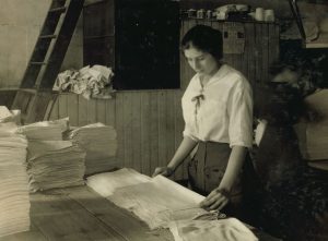 Black and white photo from 1917 of a teenage white woman surrounded by stacks of linen, looking down as she folds towels.
