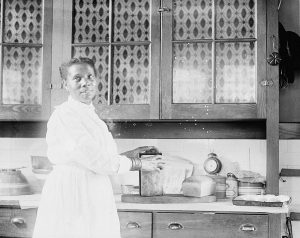 Black and white photo from 1920 of a Black woman standing in a kitchen, looking just above the camera as she lifts a freshly baked loaf of bread out of a tin.