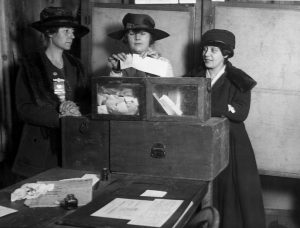 Three white women stand around a ballot box, wearing long dark coats and hats. The woman in the middle is about to place her ballot in the box.