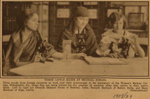 Sepia-toned newspaper photograph of three young women seated at a table looking through microscopes.