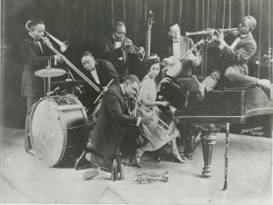 A group of six Black men kneel and stand while playing trumpets, trombones, drums, and the banjo. A Black woman sits playing a grand piano.