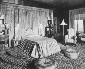A black and white archival photograph of Elaine's bedroom depicting her bed, bookcases, hassocks, and an assortment of small decorative objects.